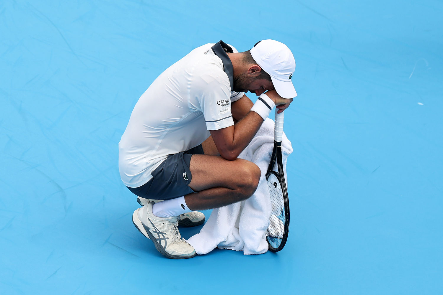 Novak Djokovic squats down and rests his head on the tennis racket he's holding, while also holding a towel (Emilee Chinn / Getty Images)